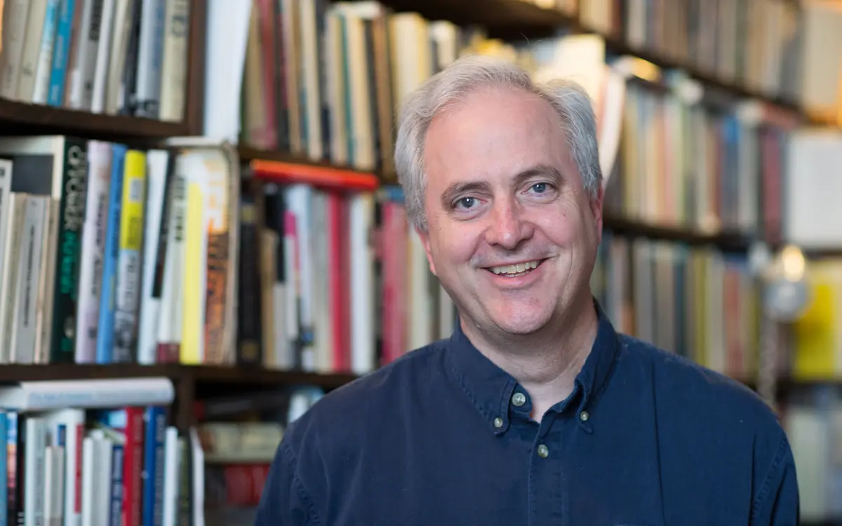 A smiling man in a blue shirt stands in front of a bookshelf filled with books.