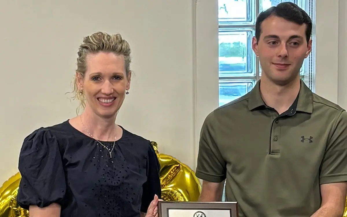 A woman in a black dress holds a plaque while standing next to a young man in a green polo shirt, both smiling in front of gold balloons.