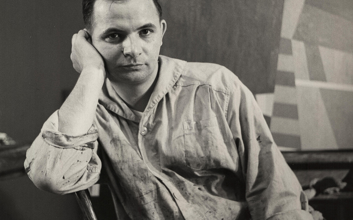 Portrait of a male artist in a studio, sitting thoughtfully with a paint-covered shirt and an abstract background.