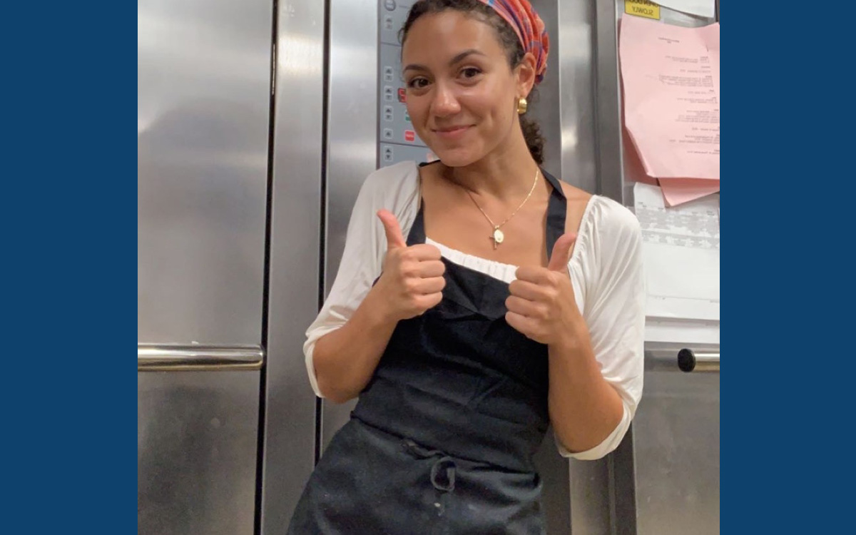 A smiling woman in a black apron and headscarf gives a thumbs-up in a kitchen setting.