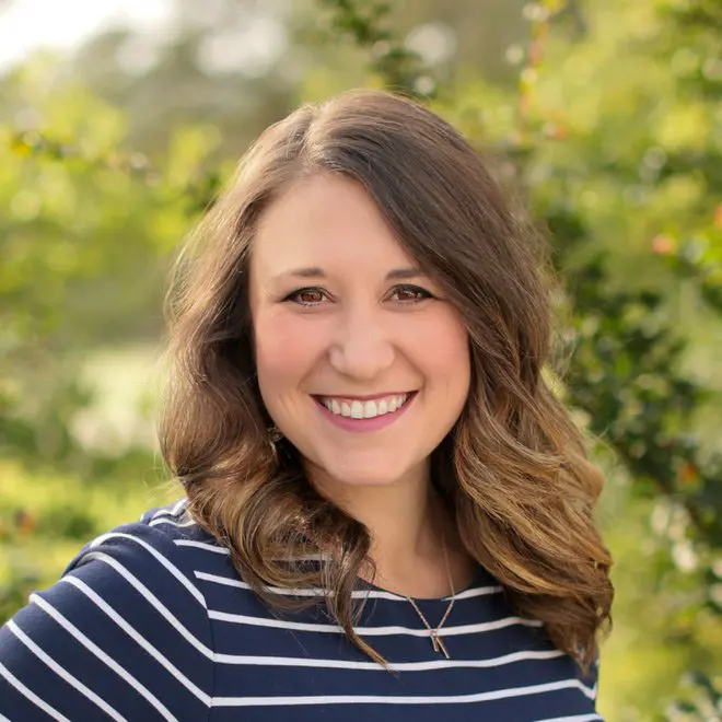 A smiling woman with long, wavy hair is posing outdoors, wearing a navy-striped shirt and surrounded by greenery.