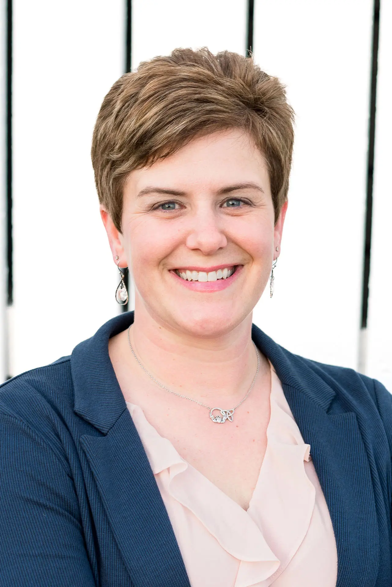 A professional headshot of a smiling woman with short, light brown hair wearing a navy blazer and a light pink blouse.