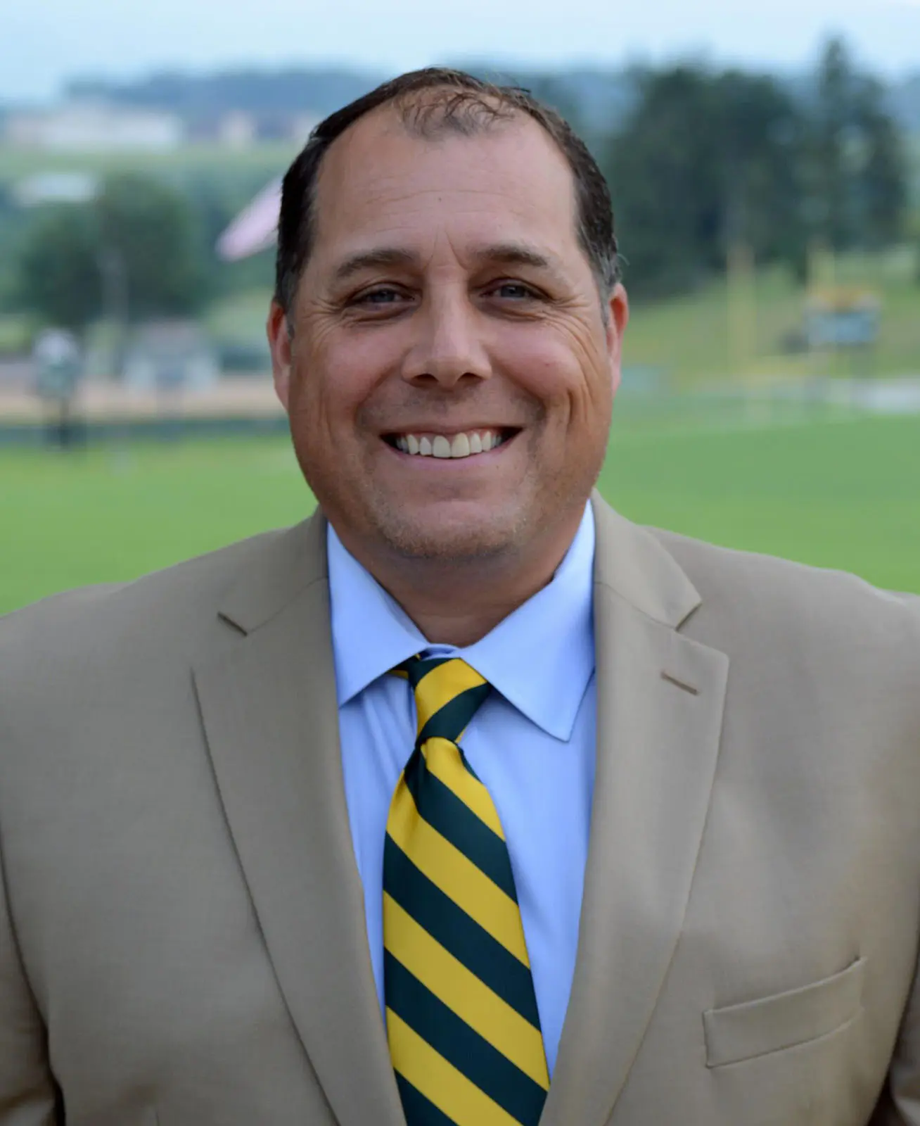 Smiling man in a suit and tie standing outdoors, with a sports field in the background.