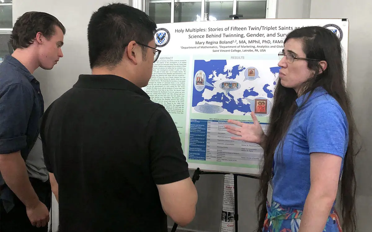 A woman presents a research poster titled "Holy Multiples: Stories of Fifteen Twin/Triplet Saints" to two attendees at a conference.