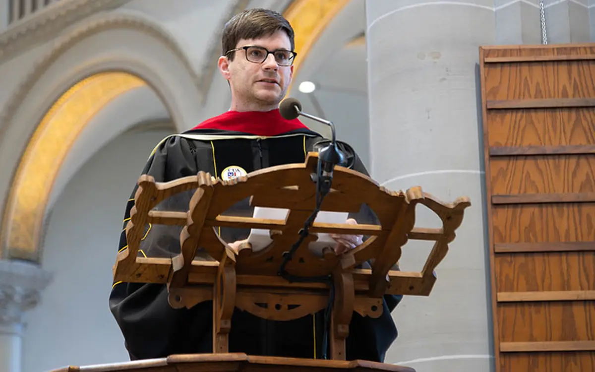 A speaker wearing a graduation gown and red hood stands at a wooden podium during a ceremony.