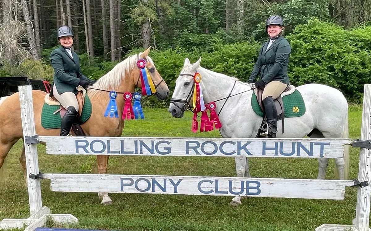 Two riders in formal equestrian attire pose with their horses, one palomino and one grey, adorned with colorful ribbons, in front of a sign for Rolling Rock Hunt Pony Club.