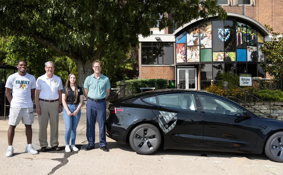 Four individuals stand beside a black Tesla in front of a modern building with large windows displaying a collage.