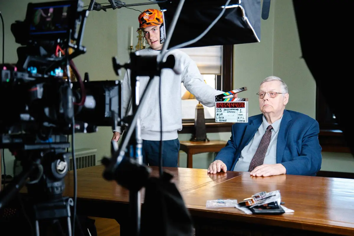 A scene from a film set featuring an older man seated at a table, looking serious, while a crew member holds a clapperboard.