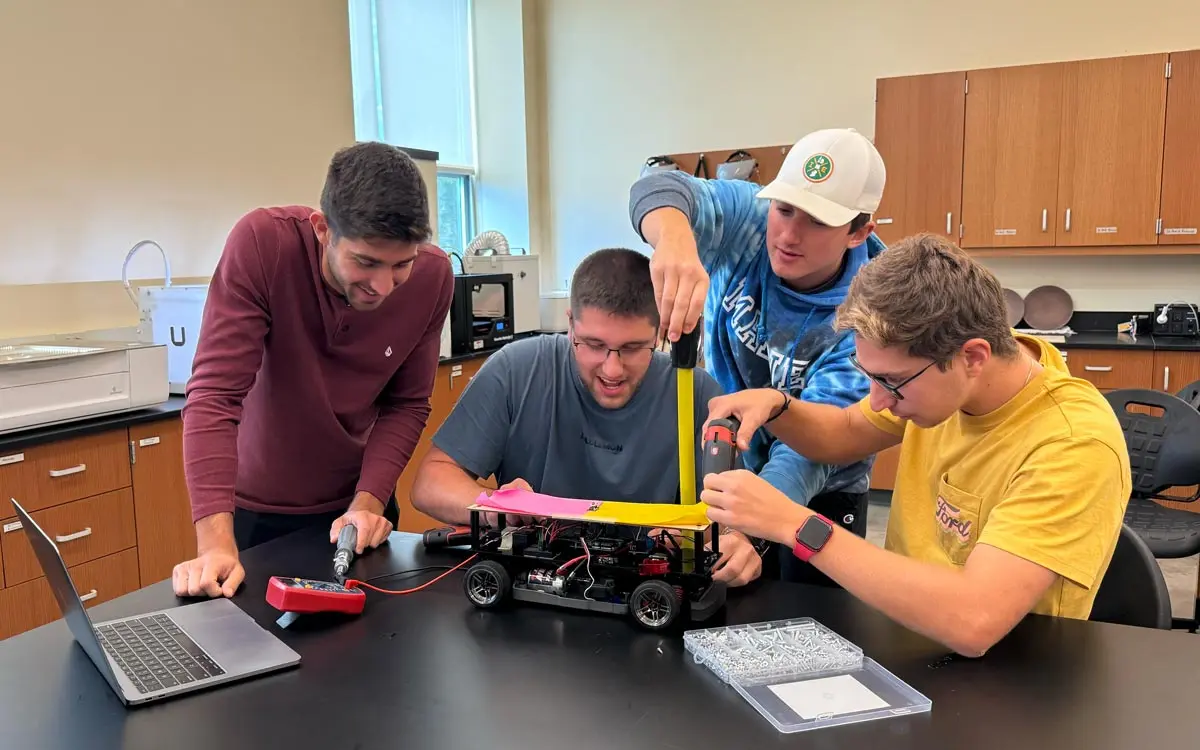 Four students collaborating on a robotics project, making adjustments to a small robotic vehicle in a laboratory setting.