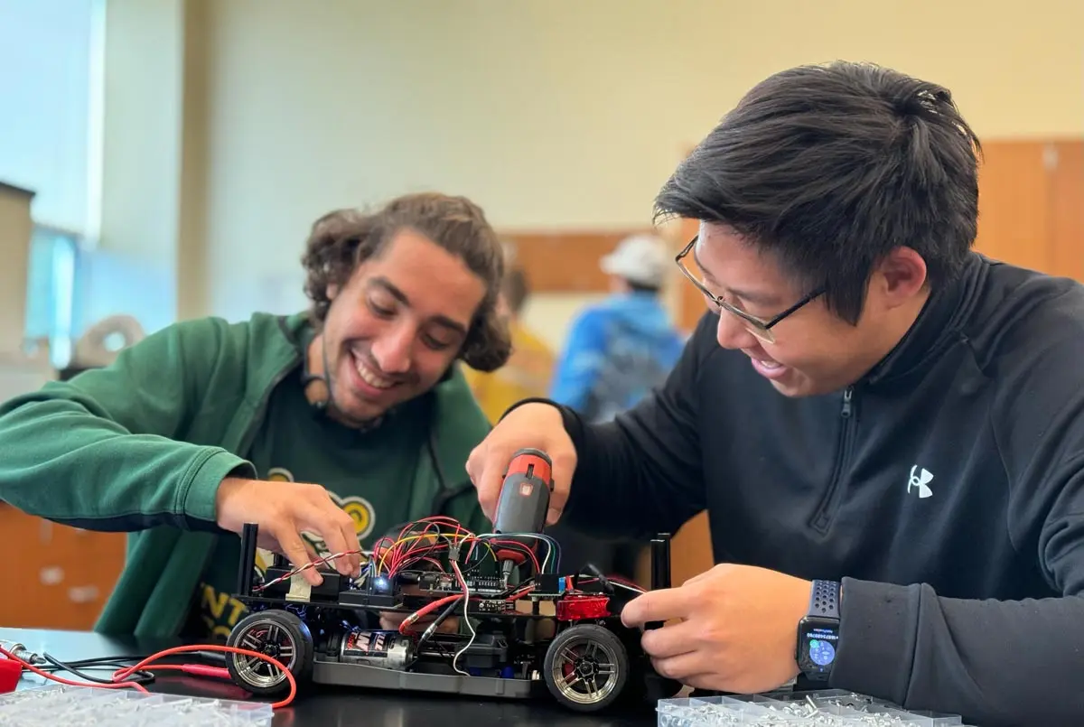 Two students collaborate on assembling a robotic car, focused on wiring and components. Their expressions reflect engagement and teamwork in a hands-on learning environment.