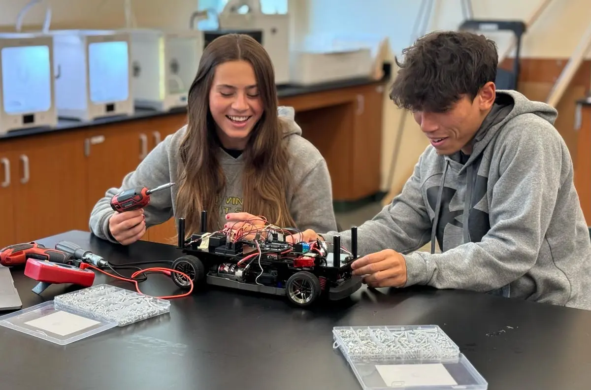 Two students collaborate on assembling a remote-controlled car, using tools and working with various electronic components in a classroom setting.