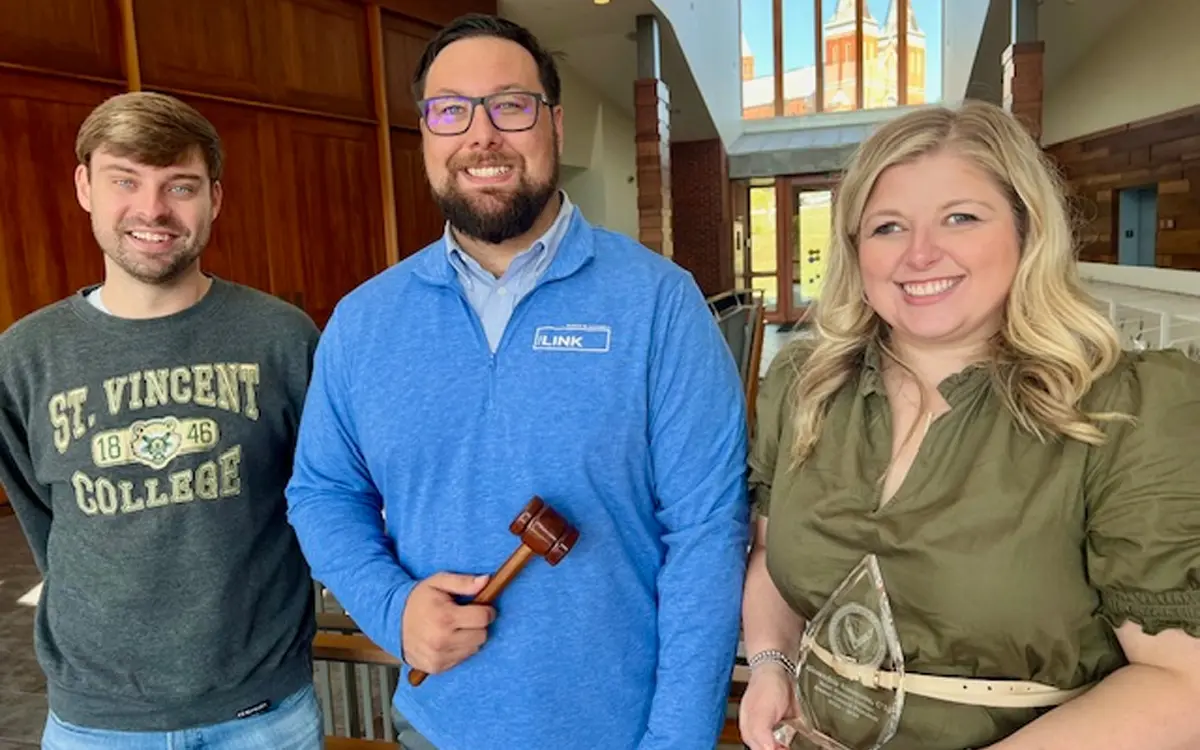 Three individuals stand together in a hallway, with one holding a gavel and another holding an award. They represent a moment of recognition and celebration.