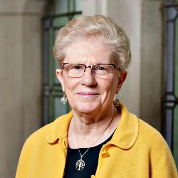 A smiling older woman with short, grey hair wearing glasses and a yellow cardigan, posing in a well-lit indoor setting.