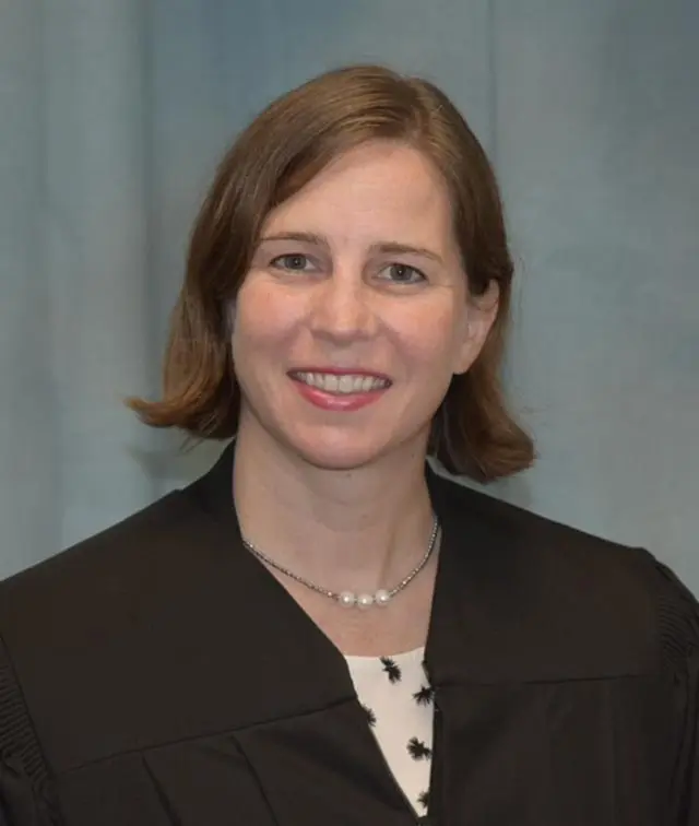 Portrait of a female judge wearing a black robe, smiling warmly against a blue backdrop.