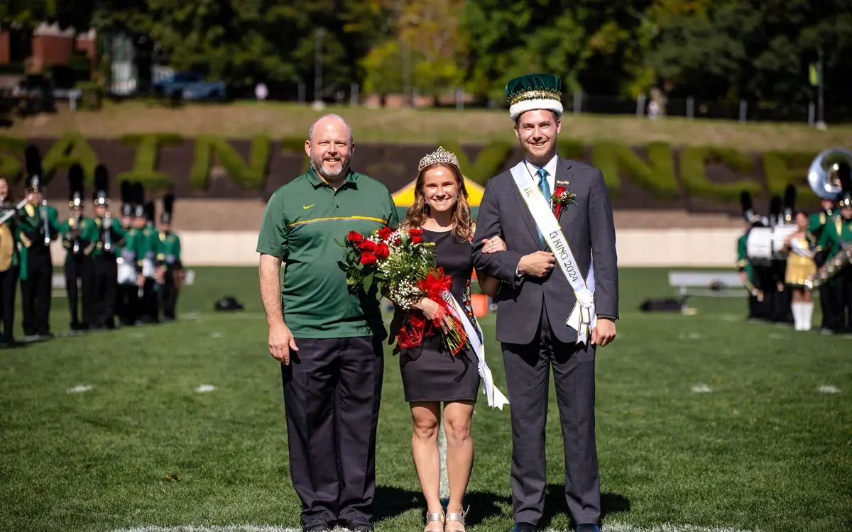 Celebration of homecoming royalty with a queen, her escort, and a king, surrounded by a marching band in the background.