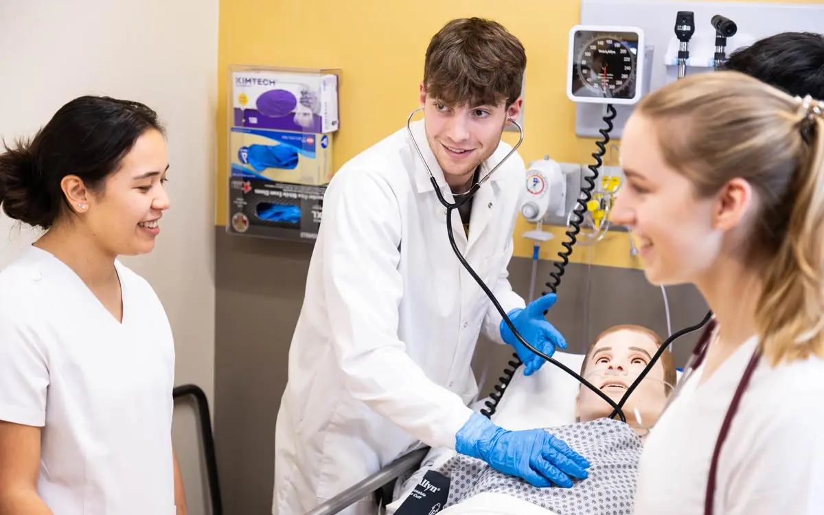 A medical student examines a patient dummy while being assisted by two colleagues in a clinical training setting.