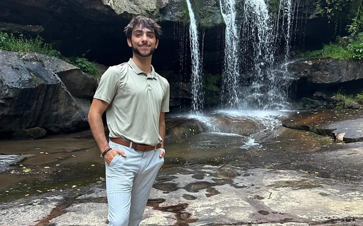 A man standing confidently by a small waterfall and rocky terrain, dressed in casual attire.