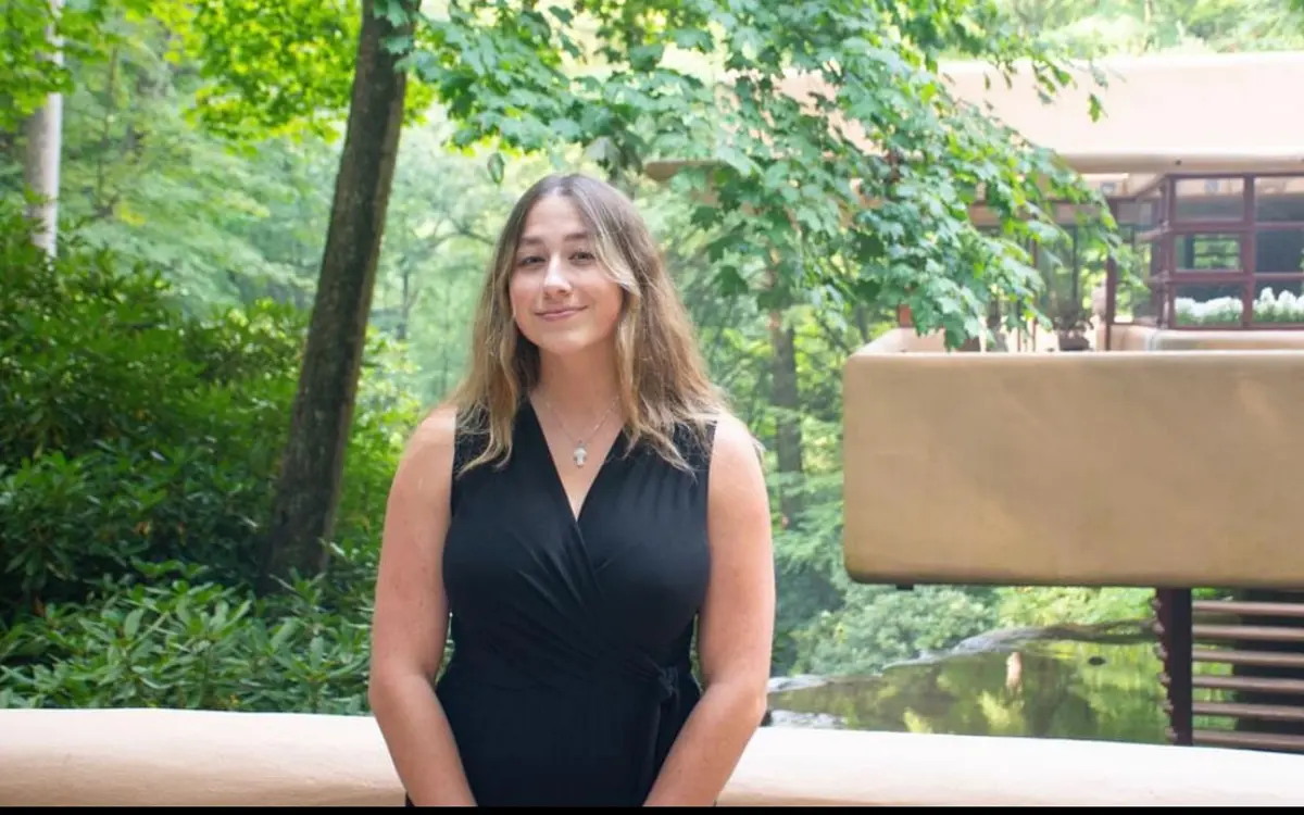 A woman in a black dress stands in front of a lush green backdrop featuring modern architecture.