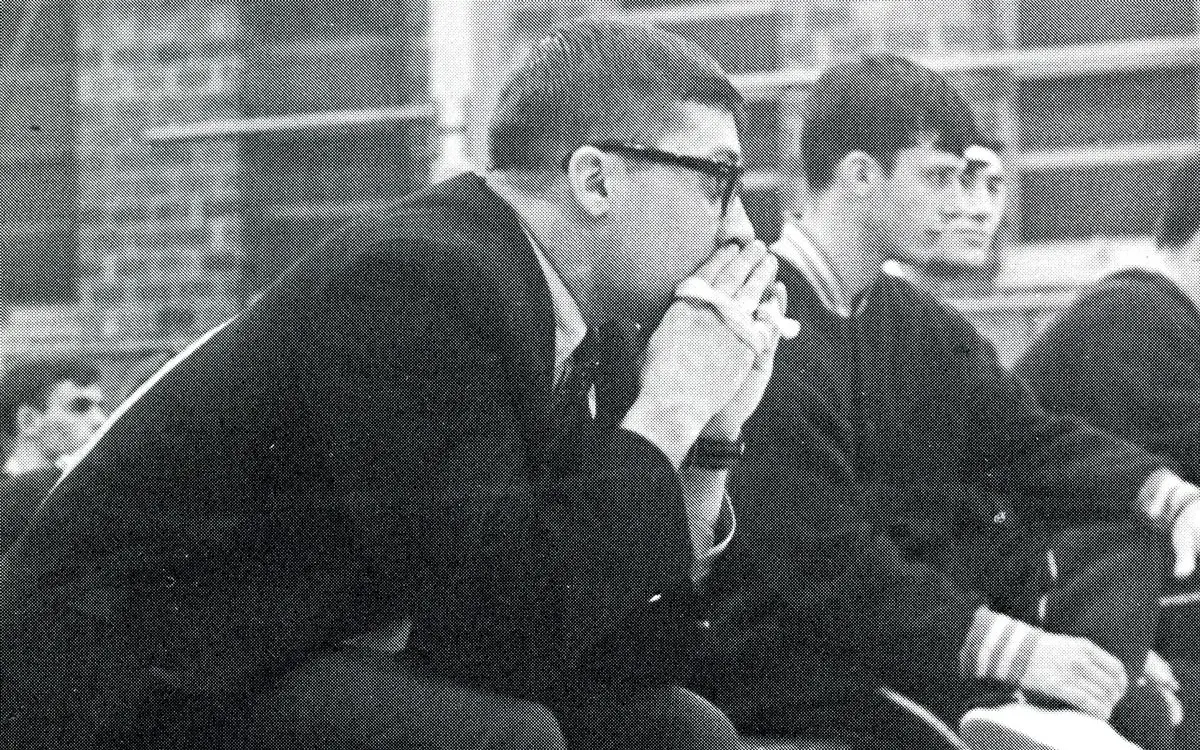 Black and white photograph of a man in glasses sitting pensively, with two young men beside him in a school setting.