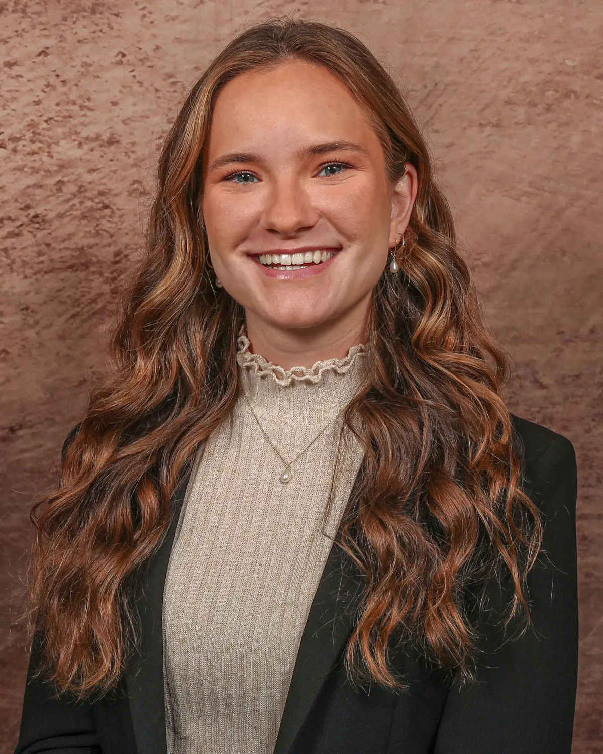 A smiling young woman with long, wavy hair, wearing a stylish black blazer and a light-colored top.