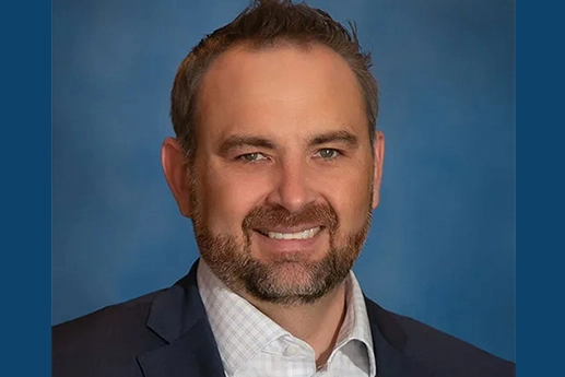 Professional headshot of a man with a beard, wearing a suit jacket and collared shirt, set against a blue background.