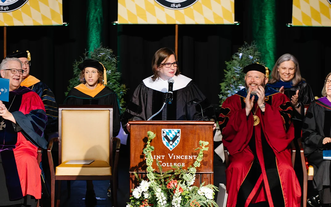 A formal graduation ceremony featuring speakers and faculty members in academic regalia, positioned at a podium with banners representing various schools.