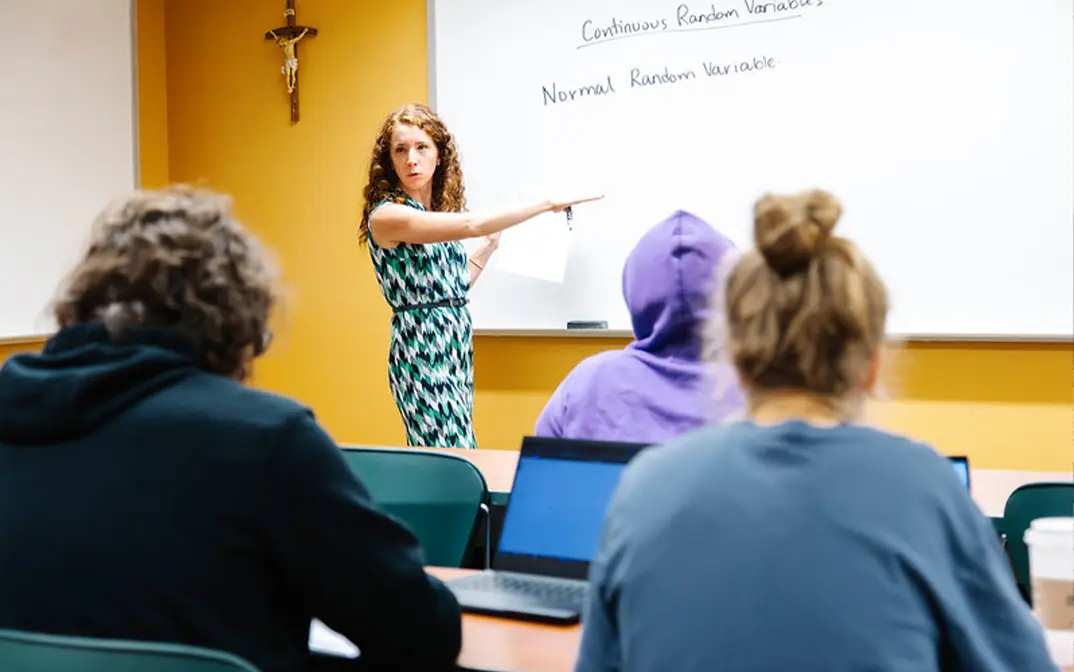 A professor points to a whiteboard while explaining concepts about random variables to a classroom of students working on laptops.