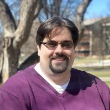 Smiling man in a purple shirt sitting outdoors with trees in the background.