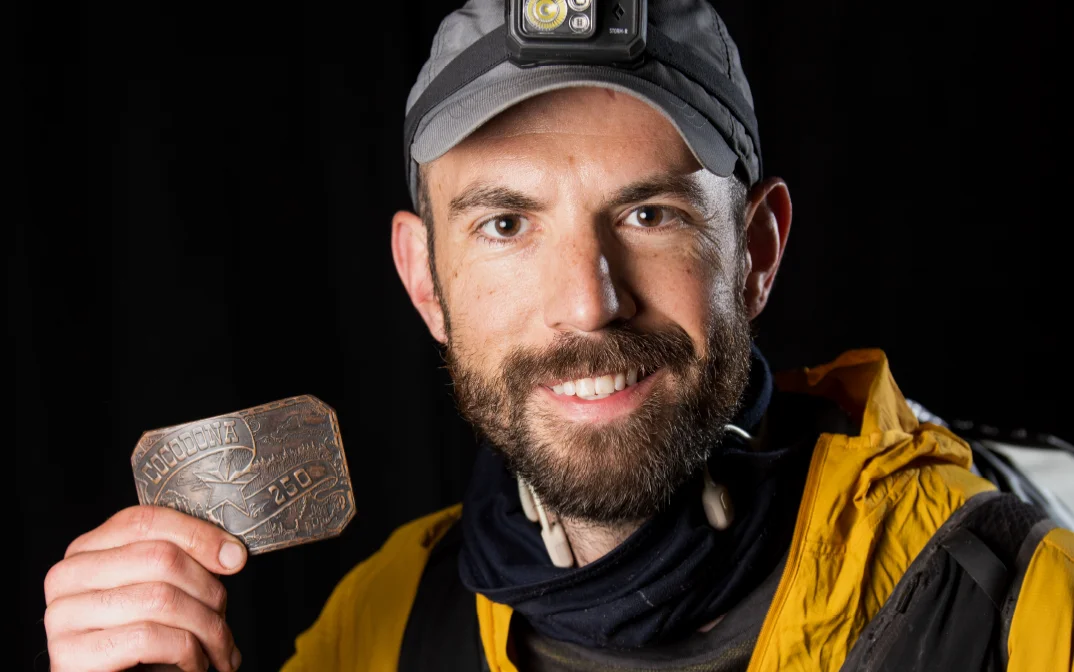 A smiling man wearing a yellow jacket and a headlamp poses with a medal in his hand.