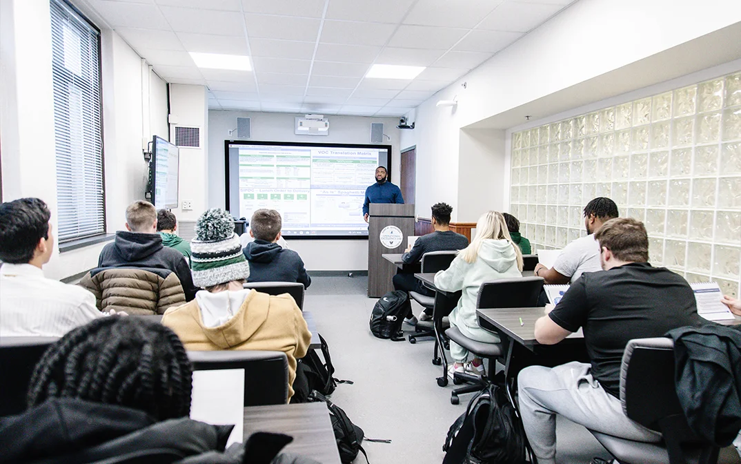 A classroom setting with a diverse group of students attentively listening to a speaker presenting information on a screen.