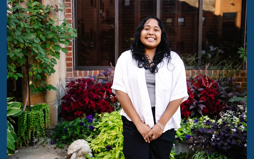 A smiling young woman stands in front of a vibrant garden filled with colorful flowers and greenery.