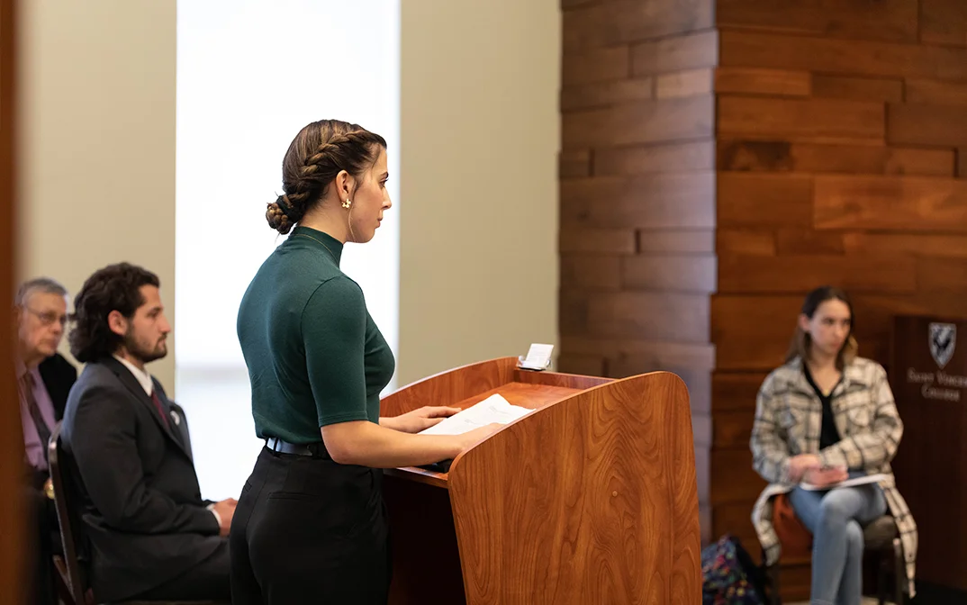 A woman in a green shirt stands at a wooden podium delivering a speech, while three individuals listen attentively in the background.