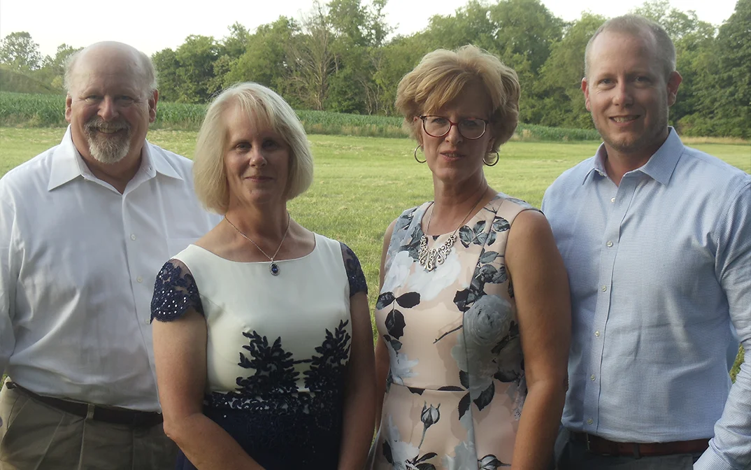 A group of four adults posing together outdoors, featuring two men and two women in formal attire, against a background of greenery.