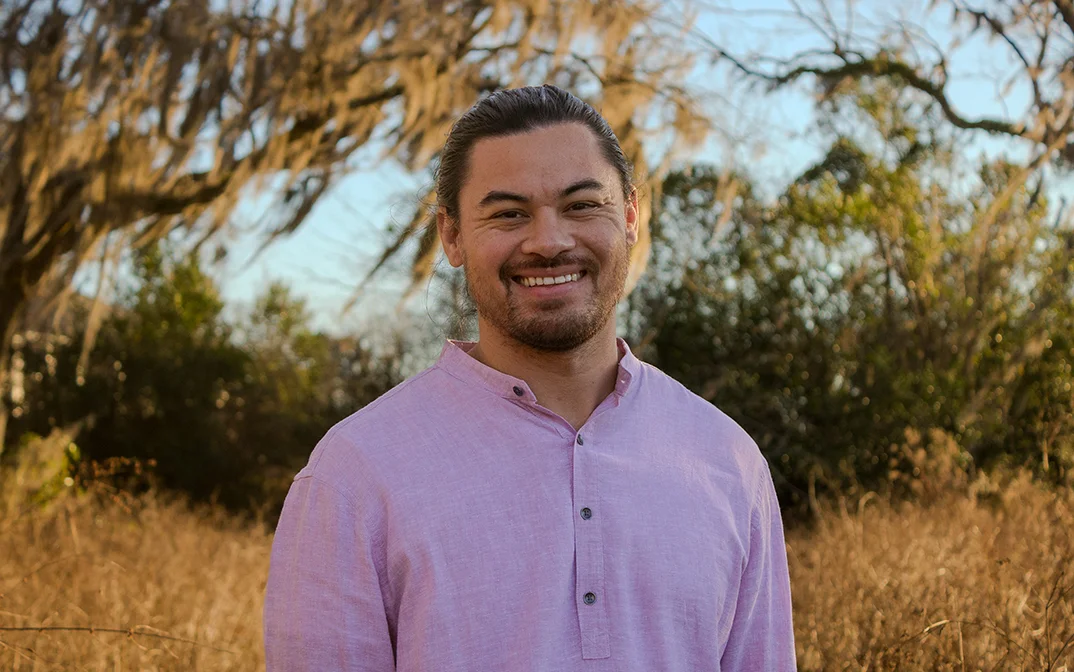 A man smiling in a natural outdoor setting with tall grass and trees in the background.