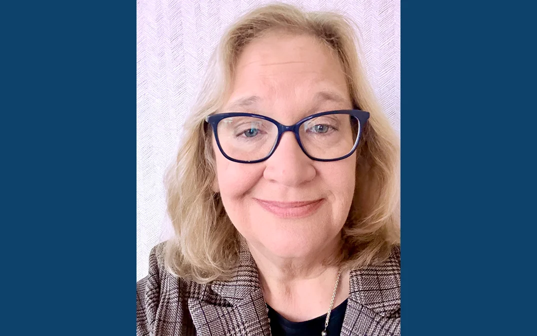 A smiling woman with blonde hair and blue glasses poses in front of a textured background.